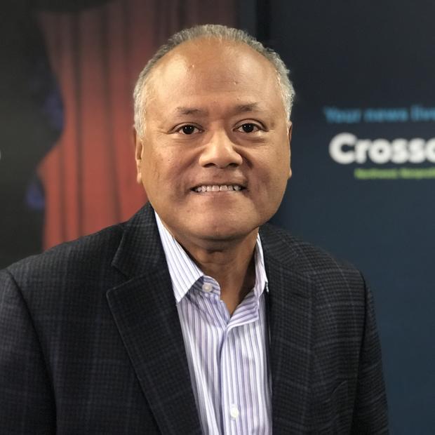 Mexican-American man with short salt & pepper hair wearing a light colored striped button-down shirt and dark suit jacket smiles at the camera. In the background are blurry posters with the Crosscut logo and unreadable text. 