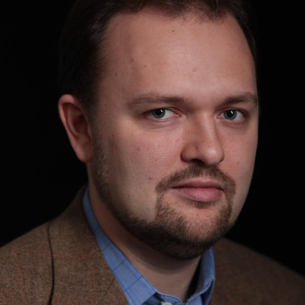 White man with dark short hair and goatee wearing a blue collared shirt and brown jacket against a dark background