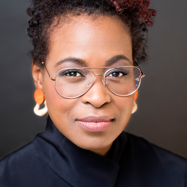 Light-skinned Black woman with short, curly, dark hear with reddish highlights wearing orange and white earrings, glasses and dark turtleneck smiles subtly at the camera