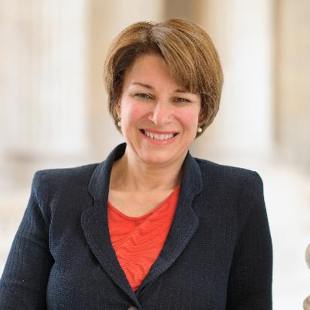 White woman with light brown hair in a short haircut wearing an orange shirt under a dark blazer smiles at the camera. In the background is a blurry building with columns. 