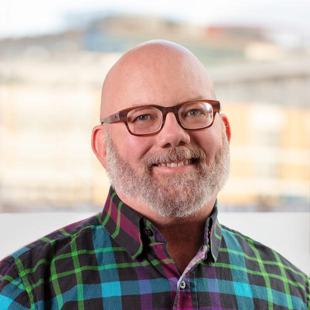 White man with greyish-white mustache and beard wearing glasses and a multi-colored checked button-down shirt against a blurry background of a building