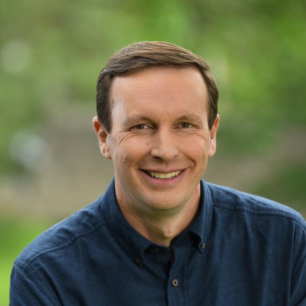 White man with brown hair wearing a deep blue button-down shirt smiles at the camera. In the background there is blurred green foliage