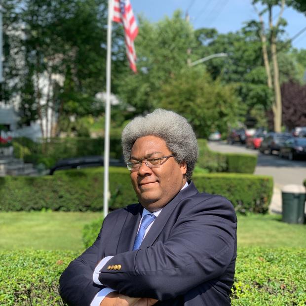 Black man with a short, grey affro wearing glasses, a light blue button-down and dark blue blazer stands with his arms crossed against an outdoor backdrop with shrubs and an American flag on a pole