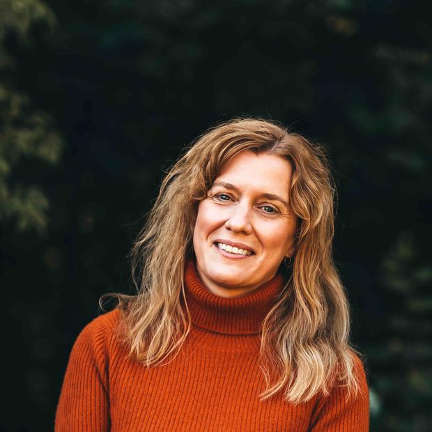 White woman with long, wavy dirty-blonde hair wearing a burnt-orange turtleneck smiles at the camera against a dark foliage background