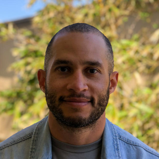 Black man with short hair, beard and mustache wearing a grey shirt under a denim shirt smiles at the camera against a background of blurred, yellow-green foliage. 