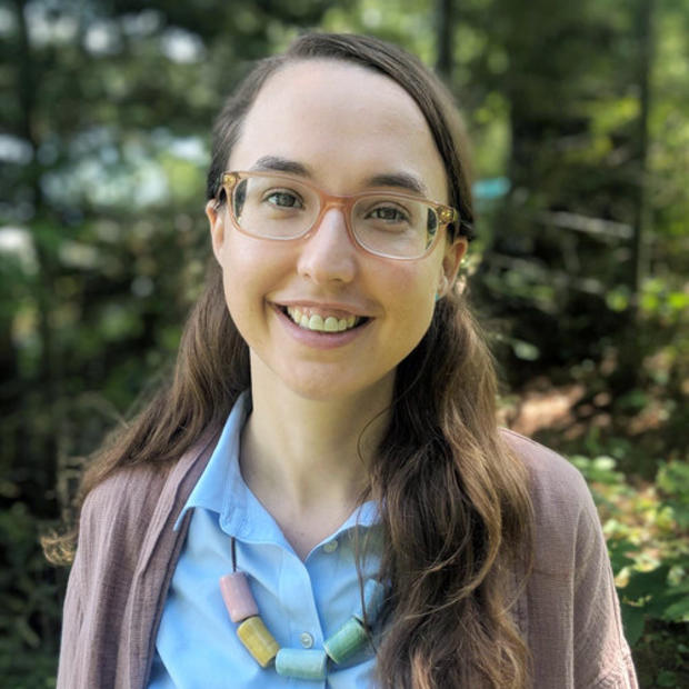 White woman with long, dark hair wearing glasses, a blue button-down shirt, a light brown cardigan and necklace with large blue and brown cylindrical beads smiles at the camera. There are trees in the background. 