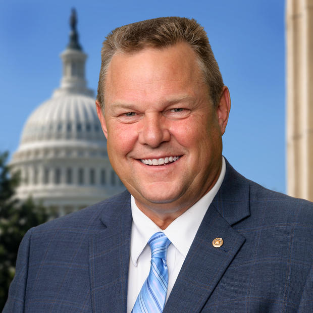 White man with brown hair in a crew cut wearing a white button-down shirt, striped, light blue tie and dark blue suit jacket with a round, gold pink on the left lapel smiles at the camera. There is  a domed building in the background. 