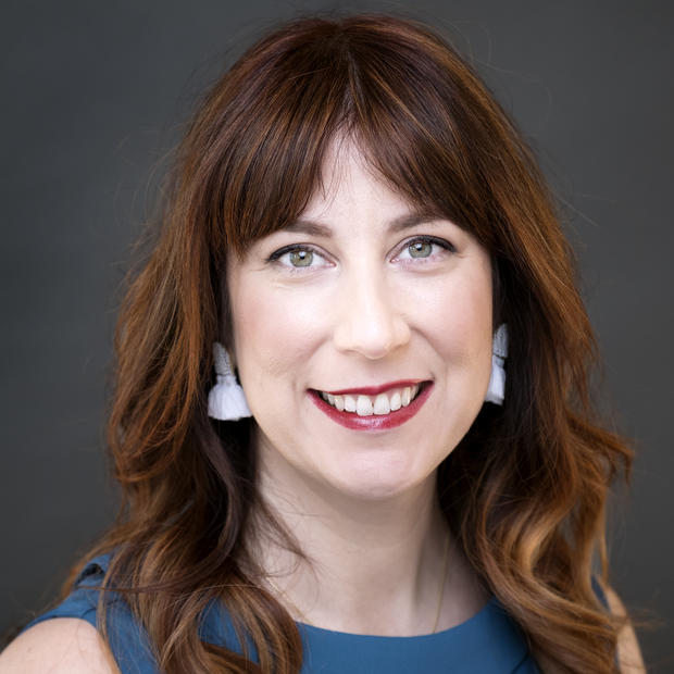 White woman with long, wavy red hair and bangs, wearing red lipstick, white earrings and a blue sleeveless shirt smiles at the camera