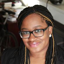 Black woman wearing glasses with long, dark braids with a few blonde braids in front smiles at the camera. She is wearing a dark blazer and her background is the inside of a living room