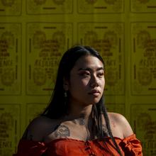 Asian woman with long, straight, dark hair wearing an orange off-the-shoulder top that reveals a tattoo on her right shoulder gazes off to the side. She is standing against a wall tiled with yellow posters of the same design