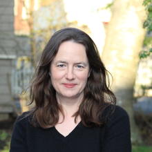 White woman with long wavy hear wearing a black v-neck 3/4 shirt stands with her arms crossed. She is outside and there is a tree and building in the background