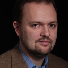 White man with dark short hair and goatee wearing a blue collared shirt and brown jacket against a dark background