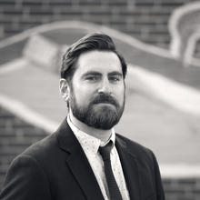 Black and white photo of a white man with thick, short, dark hair combed to the side, mustache and beard wears a white button-down shirt, dark tie and dark suit jacket. In the background is a brick wall painted with a design.  