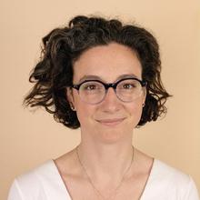 White woman with short, wavy dark hair wearing glasses, a silver pendant necklace and white top smiles at the camera against a tan background