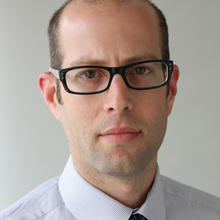 White man with dark, short hair wearing glasses and a white collared button-down shirt looks at the camera with a serious expression against a light grey background