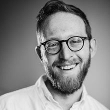 Black and white photo of a white man with short dark hair, mustache and beard wearing round-frame glasses and a white button-down shirt smiling at the camera