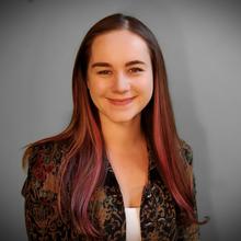 White woman with long, straight, auburn hair wearing a white shirt and dark blazer smiles at the camera against a light grey background 