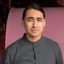 Brown-skinned man with short, dark hair wearing a grey button-down shirt stand with his hands behind his back against a pink background