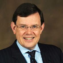 White man with dark short hair wearing glasses, a blue collared shirt, dark tie and dark jacket against a dark brown background