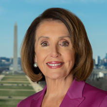 Older white woman with short, brown hear in a bob style cut wearing silver earrings, a white shirt and dark pink blazer against the backdrop of the National Mall in Washington, DC. The Washington memorial is visible. 