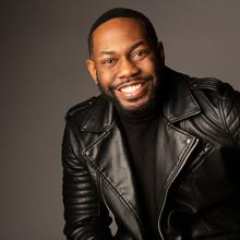 Black man with short dark hair and facial hair is wearing a black leather jacket over a black top and smiles at the camera against a dark grey background