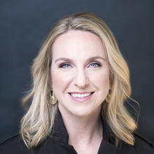 White woman with shoulder-length blonde hair wearing gold earrings, a black v-neck shirt smiles at the camera against and dark blue backdrop