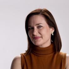 White woman with shoulder-length, straight auburn hair wearing a burnt-orange sleeveless turtleneck and gold bracelets has her arms crossed and smiles at the camera against a light grey background.