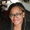 Black woman wearing glasses with long, dark braids with a few blonde braids in front smiles at the camera. She is wearing a dark blazer and her background is the inside of a living room