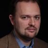 White man with dark short hair and goatee wearing a blue collared shirt and brown jacket against a dark background