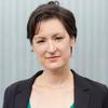 White woman with straight, short dark hair wearing a blue top and dark suit jacket stands in front of a corrugated metal wall