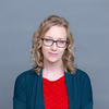 White woman with shoulder length wavy hear wearing glasses, a red shirt with a geometric design and navy blue cardigan against light grey background