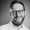 Black and white photo of a white man with short dark hair, mustache and beard wearing round-frame glasses and a white button-down shirt smiling at the camera