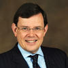 White man with dark short hair wearing glasses, a blue collared shirt, dark tie and dark jacket against a dark brown background