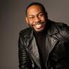 Black man with short dark hair and facial hair is wearing a black leather jacket over a black top and smiles at the camera against a dark grey background