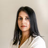 Brown-skinned woman with dark, straight hair past her shoulders wearing a gold necklace, white button-down shirt and gold bracelets has her arms crossed and looks at the camera, standing against a white background