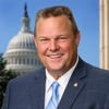 White man with brown hair in a crew cut wearing a white button-down shirt, striped, light blue tie and dark blue suit jacket with a round, gold pink on the left lapel smiles at the camera. There is  a domed building in the background. 