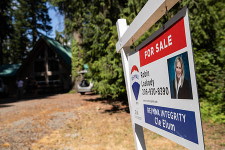 A for sale sign with an A-frame home among trees in the background