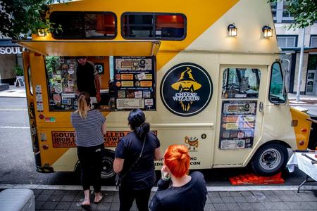 People line up on a sidewalk in front of a yellow food truck called Cheese Wizards, which sells grilled cheese sandwiches.