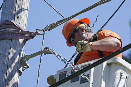 A utility worker wearing an orange shirt and helmet installs fiber cable on a utility pole while standing in a lift bucket.