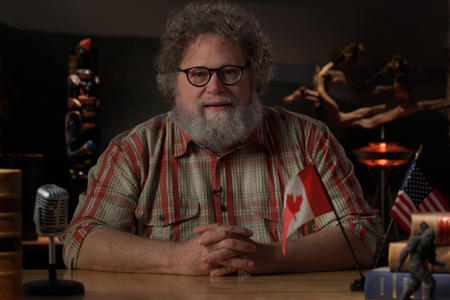 Knute Berger sitting at a table wearing a plaid shirt.  A Canadian flag and a U.S. flag are in the foreground.